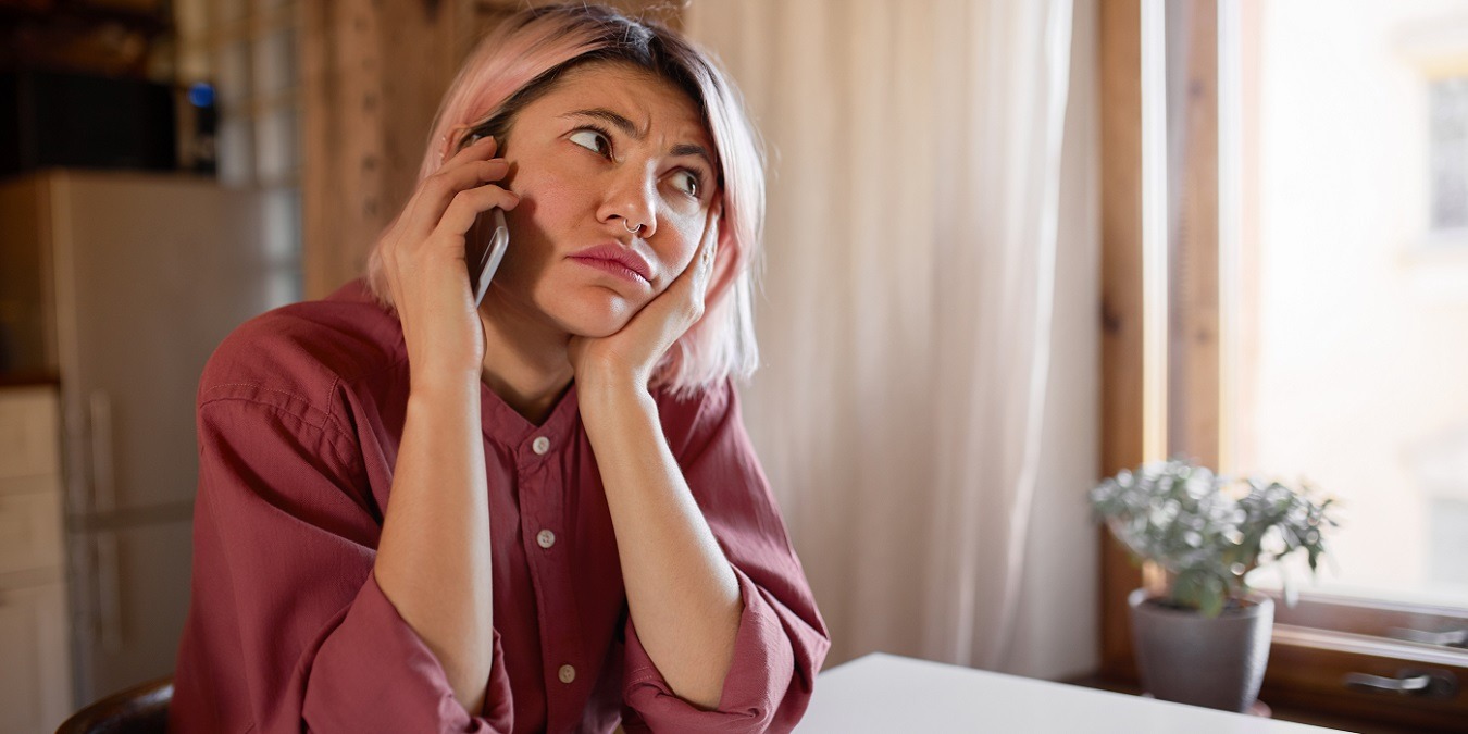Beautiful European Girl With Nose Ring And Pink Hair Sitting At Table By Window Having Bored Depressed Look, Has To Stay Home Because Of Quarantine, Speaking On Smart Phone With Her Boyfriend