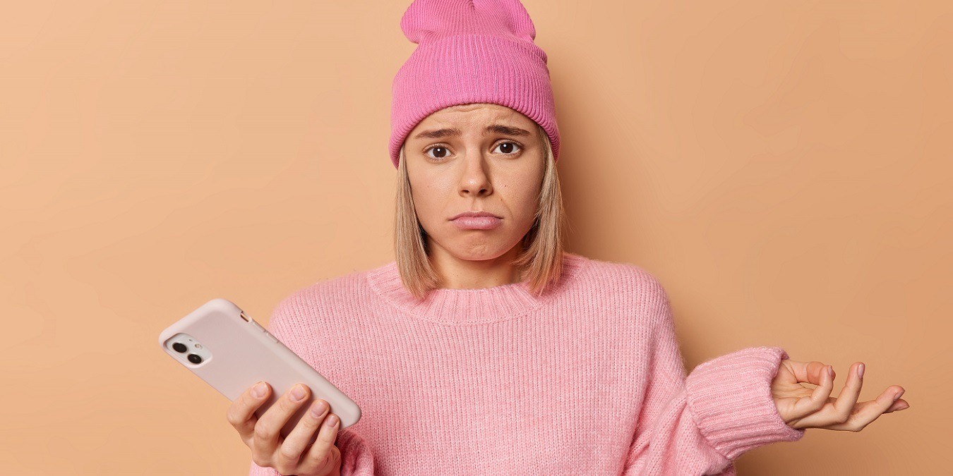 Hesitant Confused Displeased Woman Shrugs Shoulders Looks Frustrated At Camera Doesnt Know How To Make Payments Online Scrolls Social Media Wears Pink Hat And Jumper Isolated Over Brownn Background.
