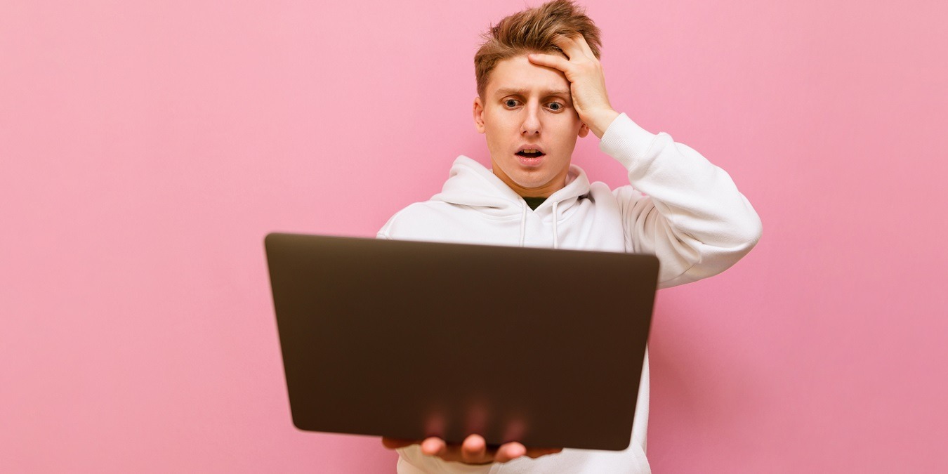 Shocked Young Man In Casual Clothes Stands With Laptop In Hands On Pink Background And Looks At Computer Screen With Surprised Face. Surprised Student Uses A Laptop, Isolated On A Pink Background. Copy Space