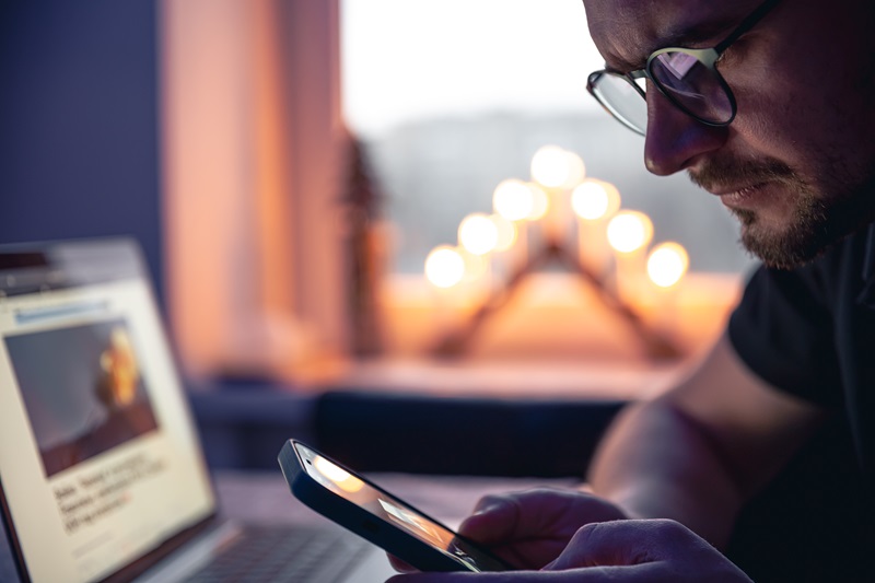 A Man With A Smartphone Sits In Front Of A Laptop Late At Night.