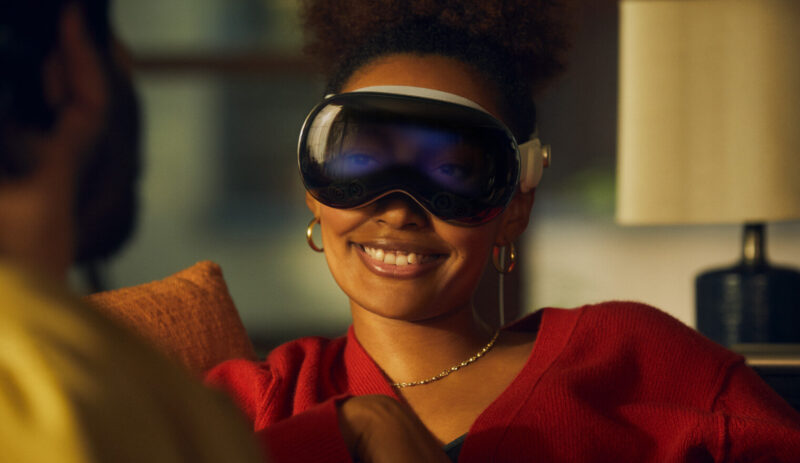 Woman smiling wearing the Apple Vision Pro in passthrough mode. You can see her eyes on the outer screen of the device.