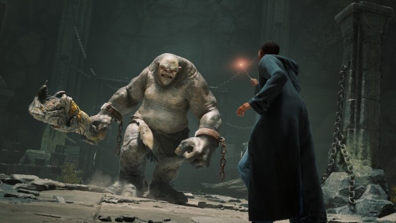 A screenshot from the game Hogwarts Legacy, showing a student wearing black robes and wielding a wand against a huge troll holding a club, charging at them.