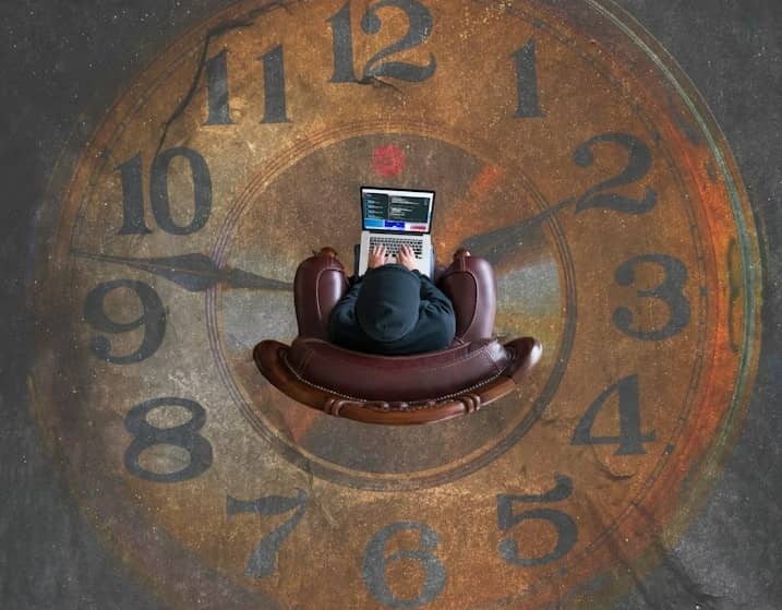 Person on laptop with a clock on the floor representing time tracking.