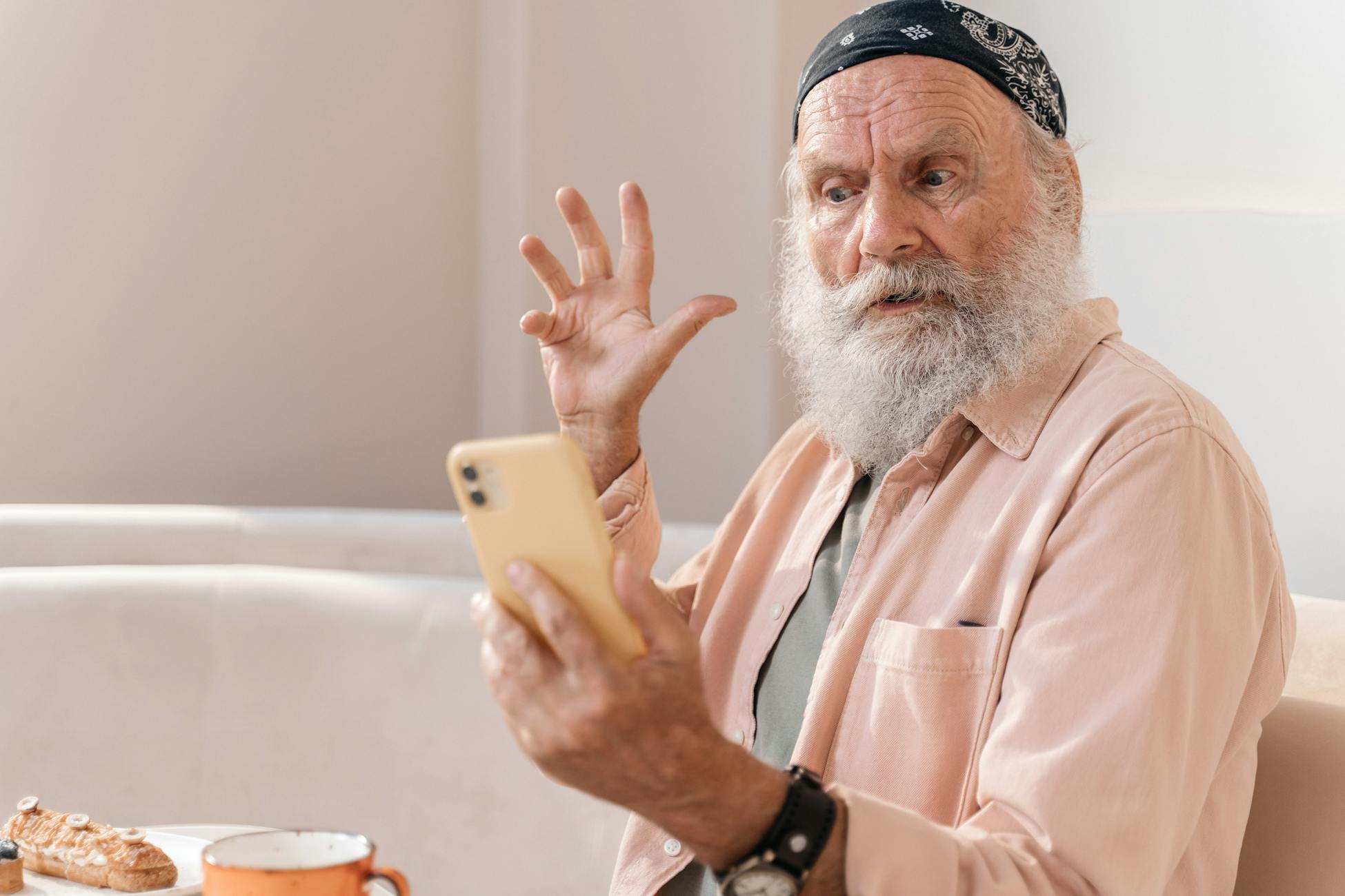 Elderly man with beard and bandana, reacting to smartphone while seated indoors.