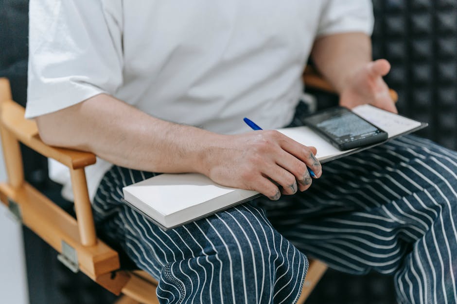 A person taking notes in a notebook while holding a smartphone, seated indoors.