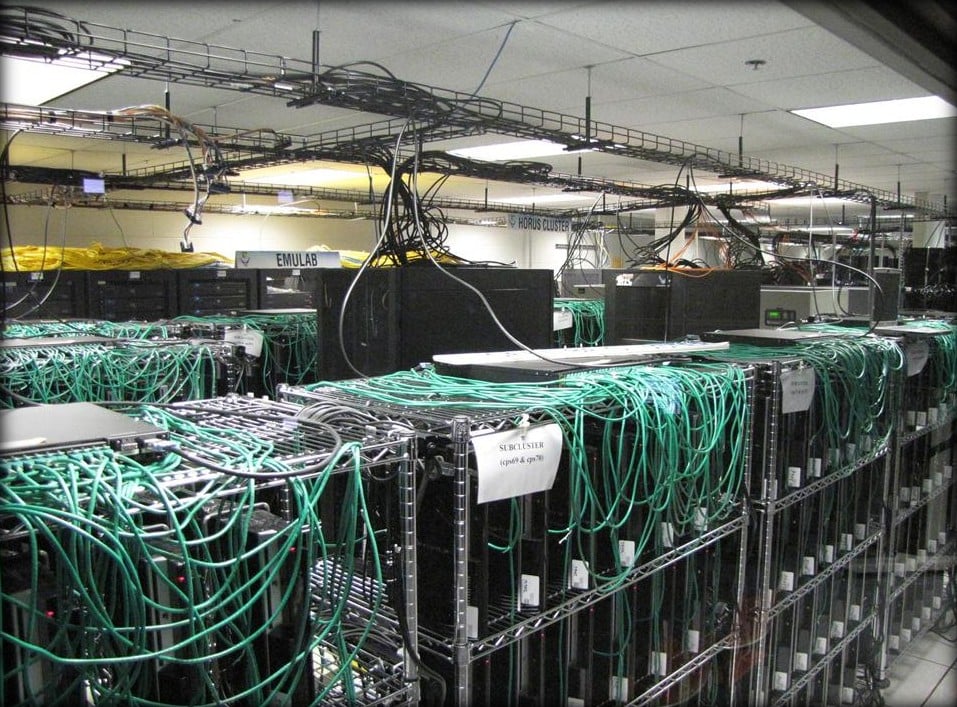 Rows of PlayStation 3 consoles on wire shelves covered in green network cables, with a sign reading Subcluster cps69 and cps70