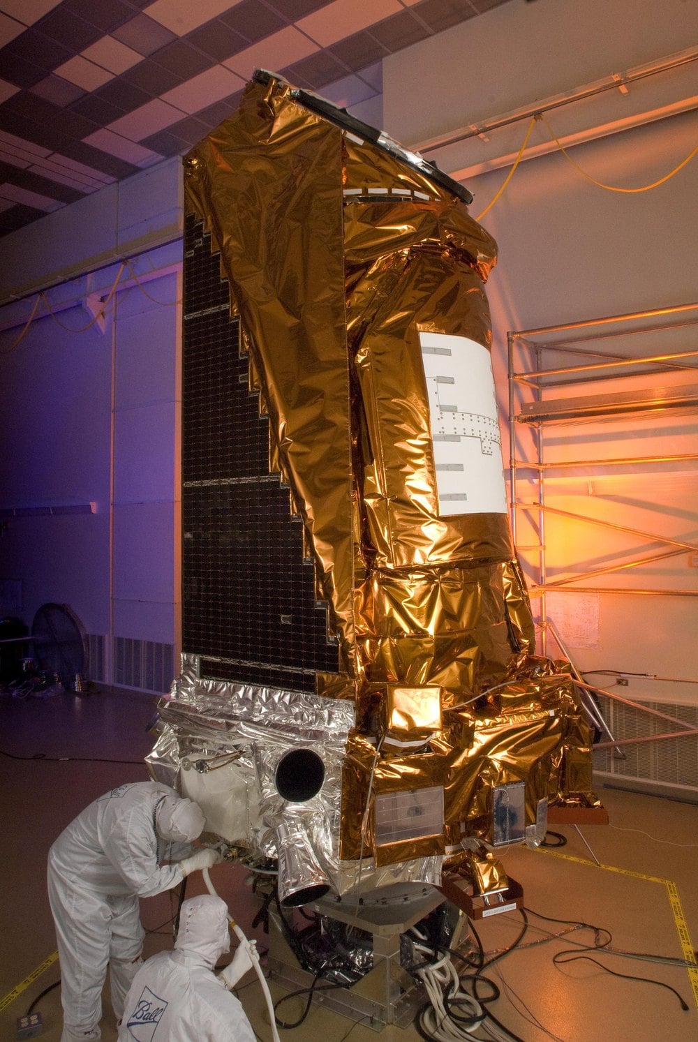 The Kepler spacecraft standing upright in a clean room at Ball Aerospace, wrapped in gold thermal blanket, with a tall flat panel of dark solar cells along one side and two technicians in white clean-room suits working at its base