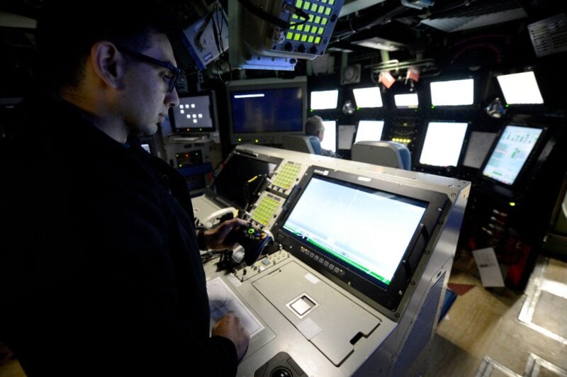 Lt. Anthony Matus holds an Xbox controller at a console aboard Pre-Commissioning Unit Colorado (SSN 788), with a large display in front of him